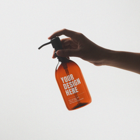 Amber dispenser bottle held by a hand with 'Your Design Here' text on a light grey background